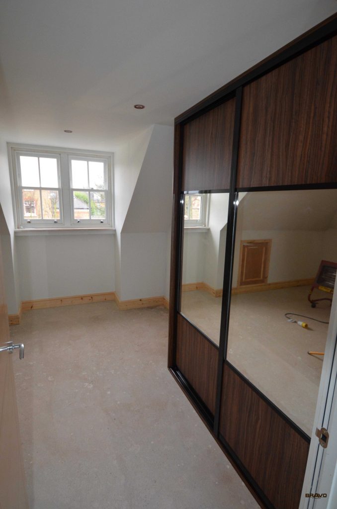 Interior of an unfinished room with a sloped ceiling, featuring large mirrored fitted wardrobes on the right and a double window. Walls are painted white and the floor is bare.