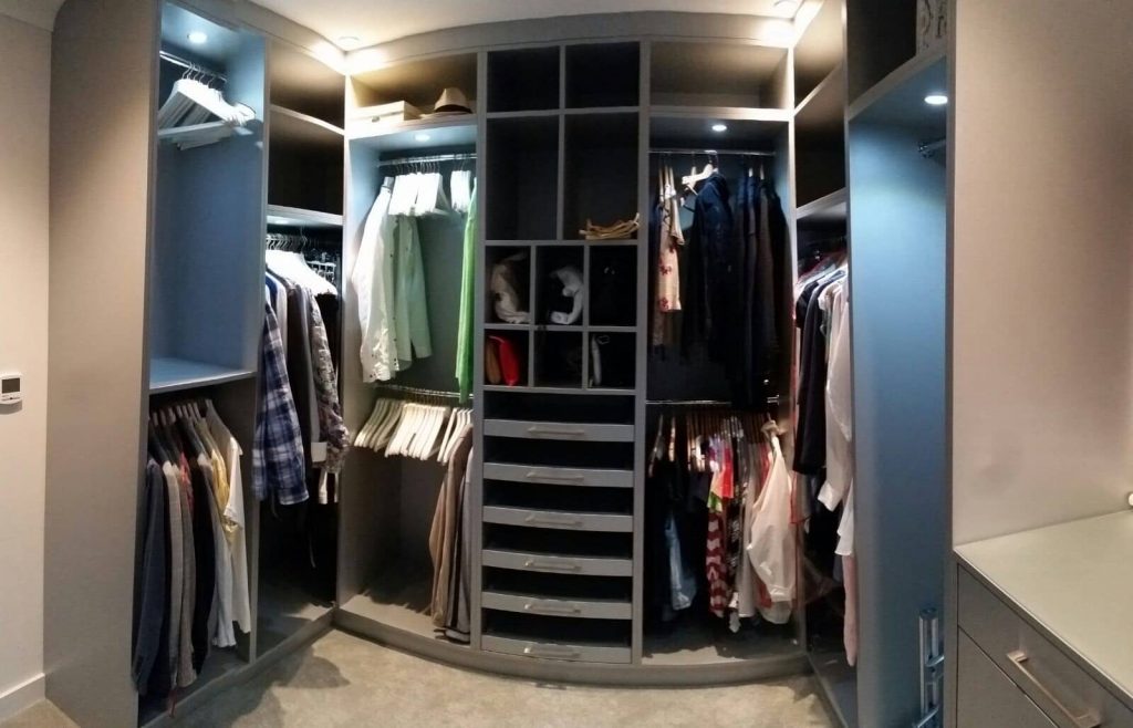 A spacious walk-in wardrobe in Mill Hill, London, with grey shelving and racks filled with various clothes and accessories. Several shirts, jackets, and pants are neatly hung, while drawers in the center contain folded items. The area is well-lit with built-in overhead lights.