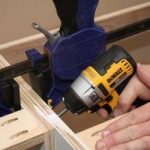 A person using a yellow and black DeWalt power drill to drive screws into a piece of wood for a walk-in-wardrobe project. The wood is held in place by a blue clamp, and there are visible pre-drilled pocket holes in the wooden boards.