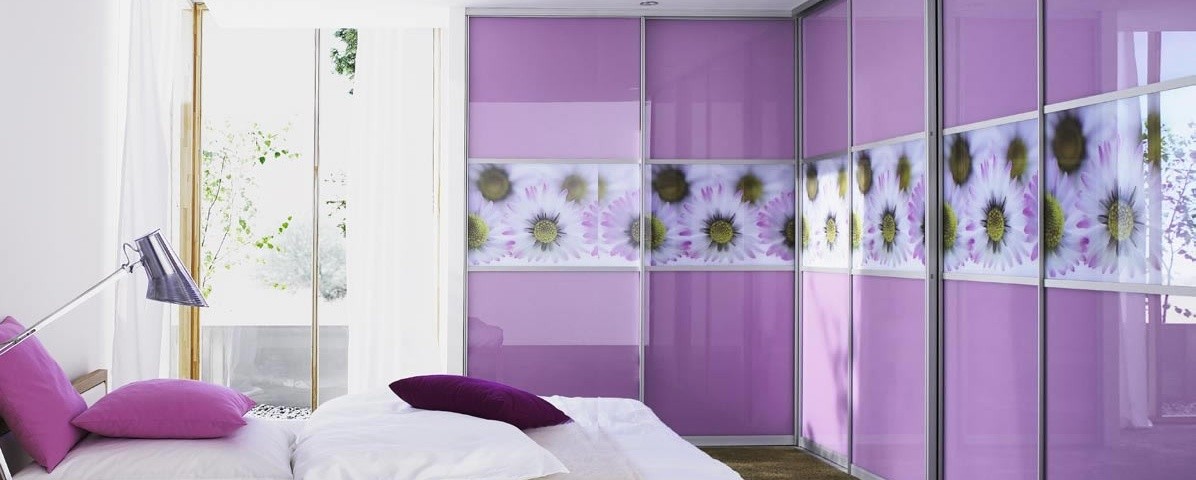 A modern bedroom featuring a large purple hinged door wardrobe with floral patterns, a white bed with purple cushions, and a bedside lamp, with an open door leading outside.