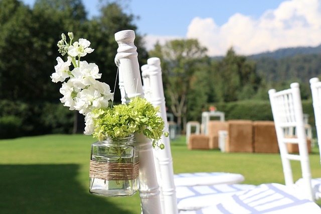 An outdoor wedding setting with white chairs, a floral arrangement in a glass jar tied to a chair, and a grassy field in the background under a clear sky.