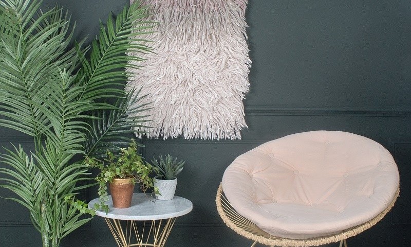 A stylish room corner with a blush papasan chair, a white marble table topped with plants, a large green fern, and a textured pink wall hanging on a dark grey wall in the fitted bedroom.