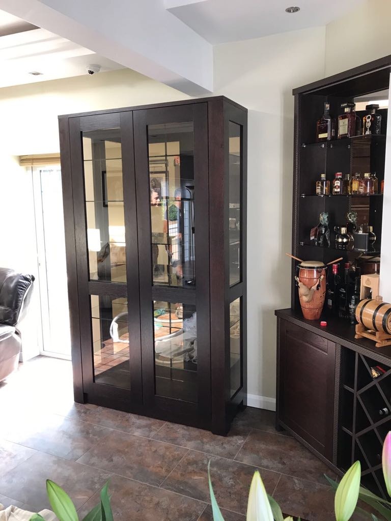 A tall dark wooden display cabinet with glass doors in a warmly lit room, positioned between a fitted wardrobe with liquor bottles and a leather chair.