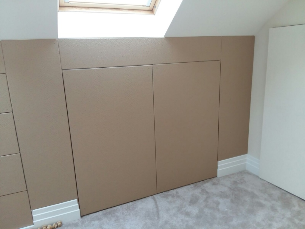 A beige room with built-in fitted wardrobes under a slanted ceiling, featuring a skylight window and a gray carpeted floor.