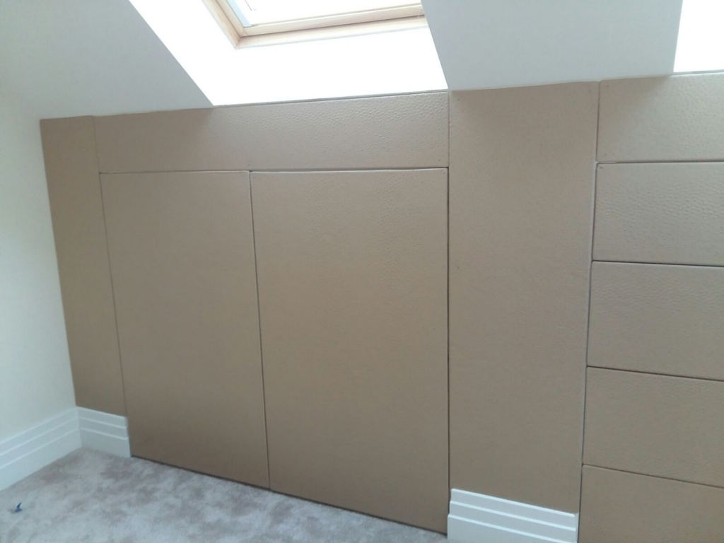 Interior view of a room with slanted walls and a skylight, featuring beige padded wall panels, a light gray carpet, and bespoke furniture.