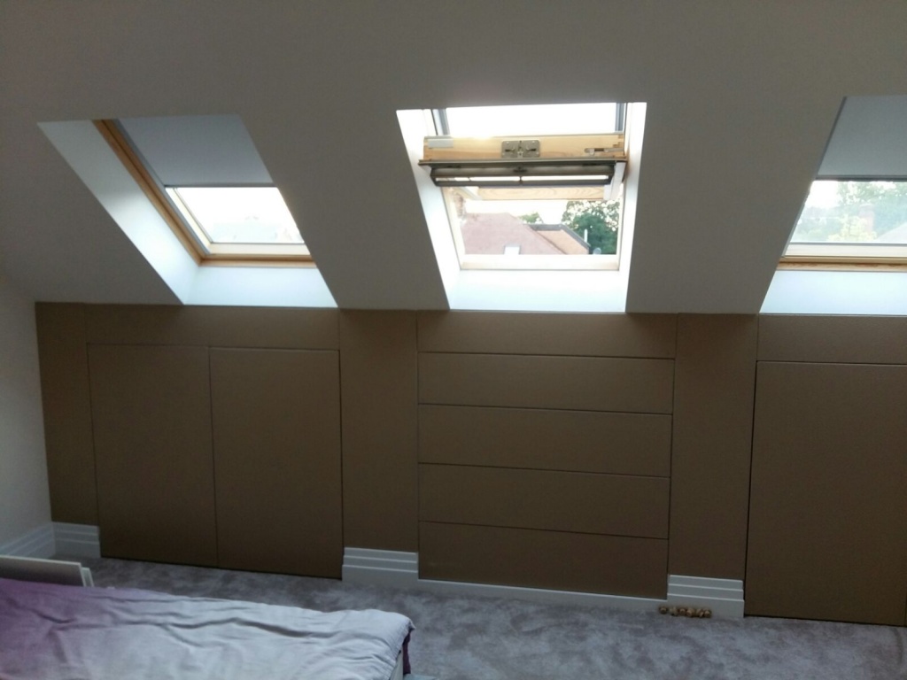 Part of the Woodside Park project in London, this room features three skylight windows on a slanted ceiling, each with a wooden frame. Below the windows are built-in storage cabinets with flush panels. The floor is carpeted, and part of a bed with a purple blanket is visible in the lower left corner.