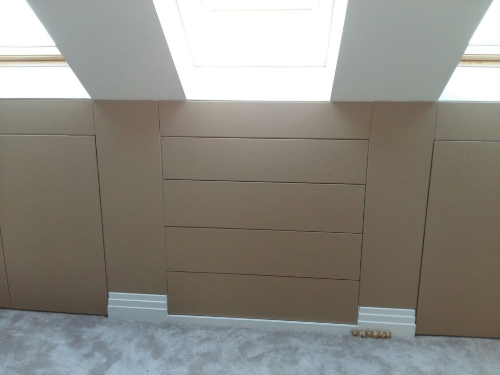 A wall with built-in storage, including two cabinets flanking a central set of four horizontal drawers. The surface is covered in a textured, light-brown material. A skylight above casts light into the space, illuminating the project. A few wooden corks lay on the light gray carpet below in Woodside Park, London.