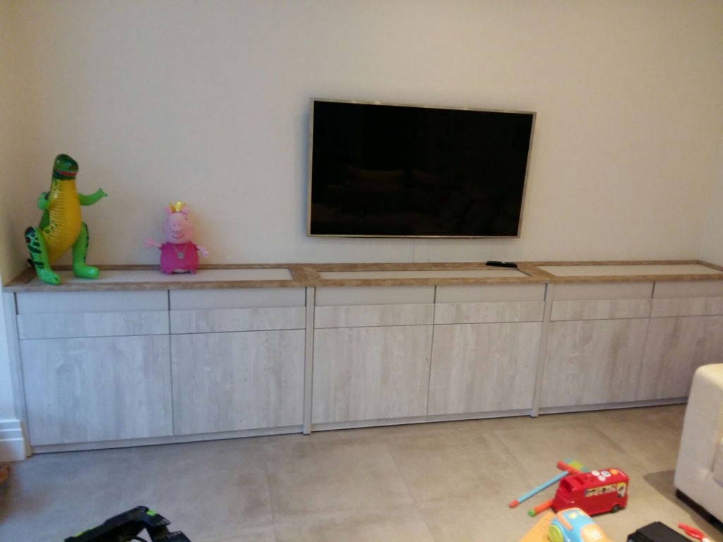 A living room featuring a wooden media console with a flat-screen TV mounted above it and fitted wardrobes along the side. On top of the console, there are two toys: a green dinosaur and a pink pig. There are scattered toys on the floor.