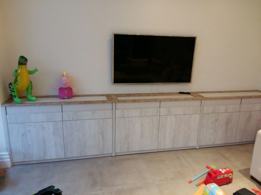 A modern living room with a large flat-screen TV mounted on the wall above a long gray wooden cabinet. Besides the cabinet, there stands a fitted bedroom wardrobe enhancing the space's functionality. On the cabinet, there are toy figures of a green dinosaur and a pink pig. Various toys are scattered on the