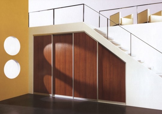A modern interior setting featuring a wooden under-stair storage cabinet with bespoke furniture elements, white stairs with a metal railing, and yellow walls with circular cutouts casting shadows.