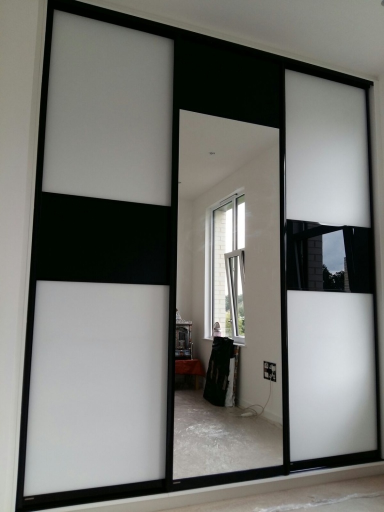 A modern sliding door with a geometric design, featuring alternating black and white panels and a central rectangular mirror, seamlessly integrates into a 3 m high wardrobe. The door opens to a room in Edgware, London, with large windows that allow natural light to enter. A partially open window and some furniture are visible inside.