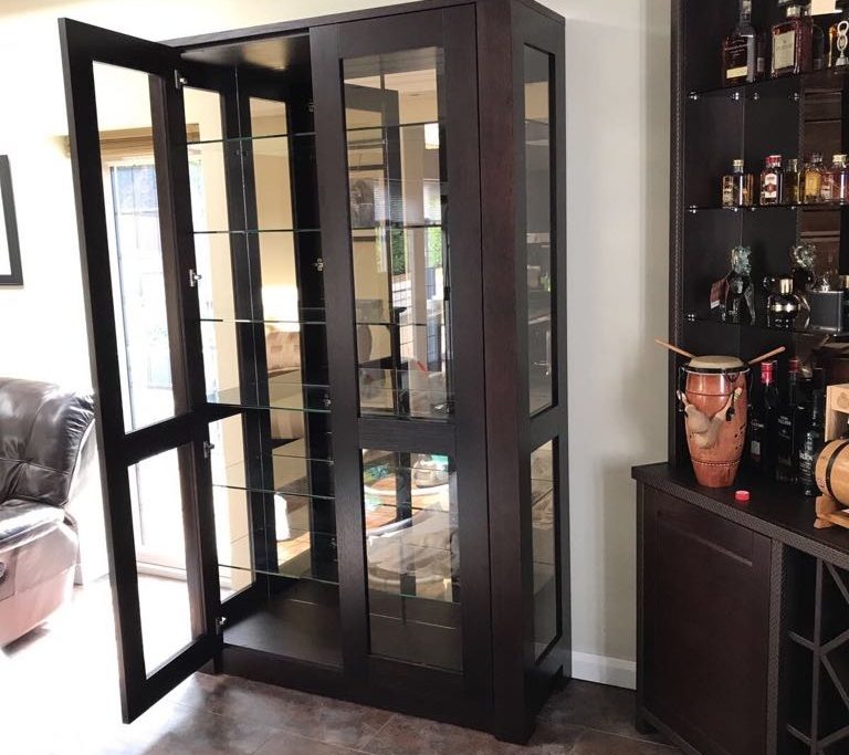 A modern living room in London features a tall, dark brown glass display cabinet with open doors. Adjacent to it is a matching shelving unit holding various bottles and a drum. The room has tile flooring, a luxurious brown leather chair, and a partial view of a window showcasing bespoke joinery.