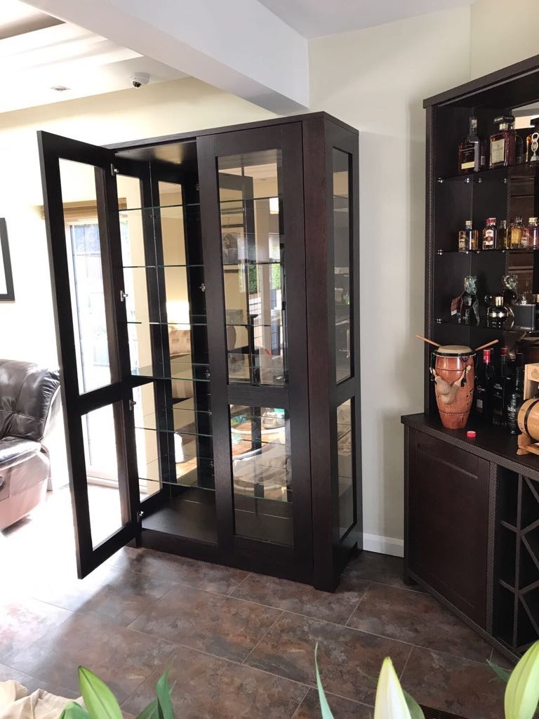 A modern living room in London features a tall, dark brown glass display cabinet with open doors. Adjacent to it is a matching shelving unit holding various bottles and a drum. The room has tile flooring, a luxurious brown leather chair, and a partial view of a window showcasing bespoke joinery.