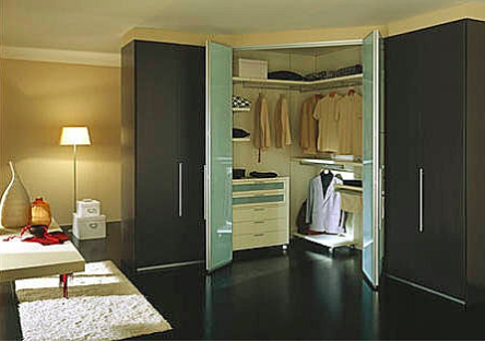 A modern, well-lit bedroom features a large, open corner fitted wardrobe with frosted glass doors. The wardrobe has hanging clothes, shelves, and drawers. Nearby, a floor lamp illuminates a small side table with boxes and a vase seated on it. A white bench is in the foreground.
