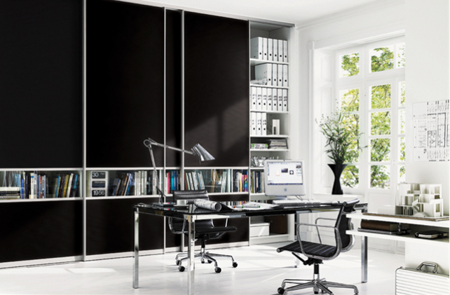 A modern home office with sleek black sliding door cabinets, open shelves with books, and binders. The room features a large glass desk with dual monitors, a keyboard, a mouse, and office supplies. A black ergonomic chair and large windows with a scenic view complete the setup.
