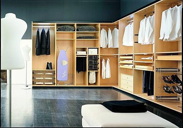 A spacious walk-in closet designed by expert fitted wardrobe manufacturers features wooden shelves and hangers. Neatly hung shirts, folded clothes, shoes, and accessories are organized throughout. An ironing board with a purple cover stands in the middle, while a white mannequin is placed to the left.