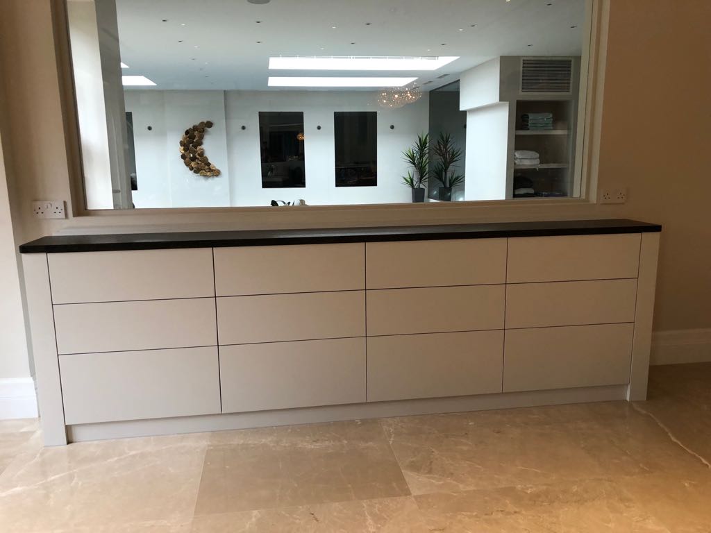 A modern kitchen featuring a large white island with sleek drawers, topped with a dark countertop, set against a backdrop of glossy cabinets and bespoke furniture. A stylish chandelier and plants add decorative touches.