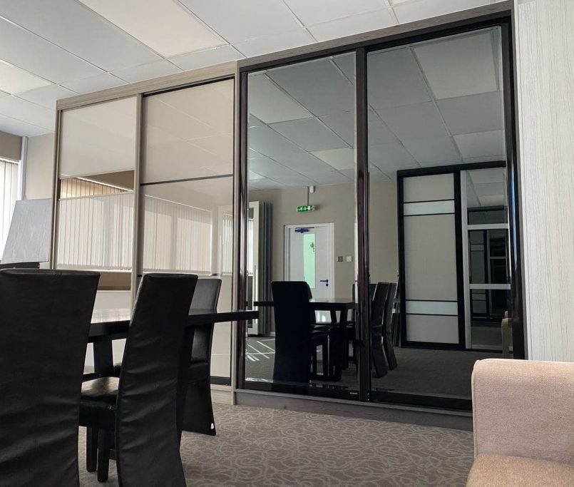 A modern conference room with a long table surrounded by black leather chairs. The room features large glass sliding doors, a carpeted floor, and a small sofa in the corner. Well-lit with natural light from a window on the left, it also includes handy sliding door wardrobe tips for organization.