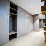 A modern, spacious walk-in closet with built-in grey cabinets and shelves made of luxury bespoke furniture under bright recessed lighting. A wooden staircase with brown steps and a black railing is visible on the right. The floor is made of light-colored wood.