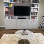 A cozy living room features a large white bookshelf with a flat-screen TV at its center, surrounded by neatly arranged books and decorative items. Below the TV is a cabinet with closed compartments. In the foreground, a round marble coffee table sits on a plush white rug. A small decorated Christmas tree is visible on the right side.