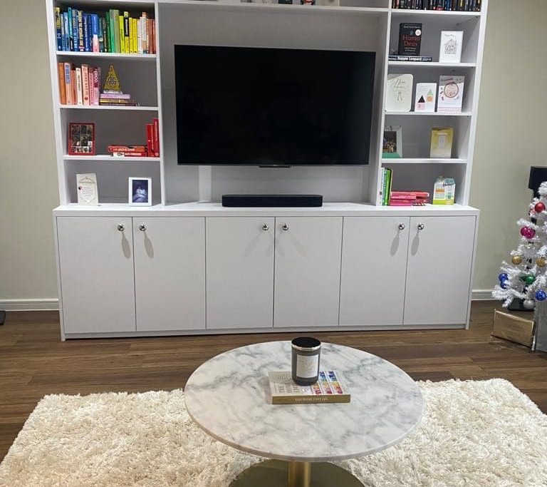 A cozy living room features a large white bookshelf with a flat-screen TV at its center, surrounded by neatly arranged books and decorative items. Below the TV is a cabinet with closed compartments. In the foreground, a round marble coffee table sits on a plush white rug. A small decorated Christmas tree is visible on the right side.