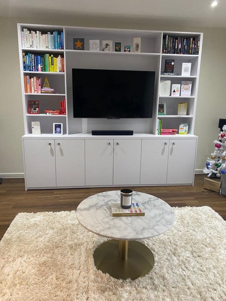 A cozy living room features a large white bookshelf with a flat-screen TV at its center, surrounded by neatly arranged books and decorative items. Below the TV is a cabinet with closed compartments. In the foreground, a round marble coffee table sits on a plush white rug. A small decorated Christmas tree is visible on the right side.