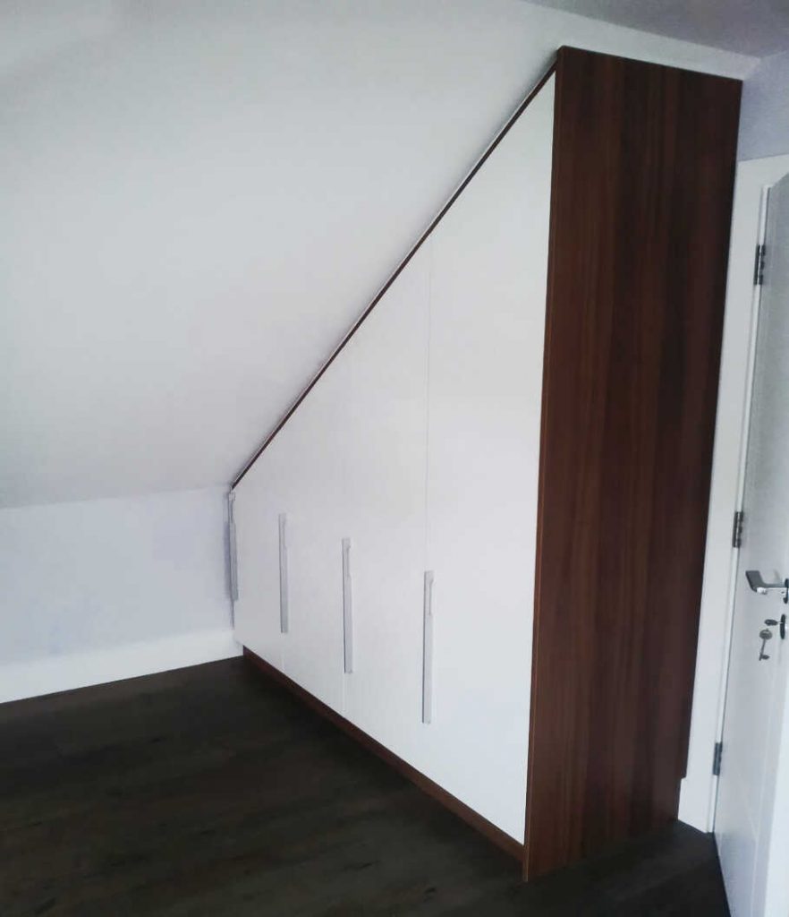 A fitted wardrobe with a sloped top aligning perfectly with the ceiling, featuring brown wood sides and elegant white front doors with silver handles. The wardrobe is positioned in the corner of a room with dark flooring and white walls, seamlessly blending into spaces with sloping ceilings.