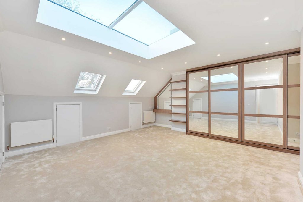 Modern spacious attic room with skylights, neutral carpet, large sliding glass doors, and a staircase leading to a lower level. Bright and airy interior featuring bespoke furniture.