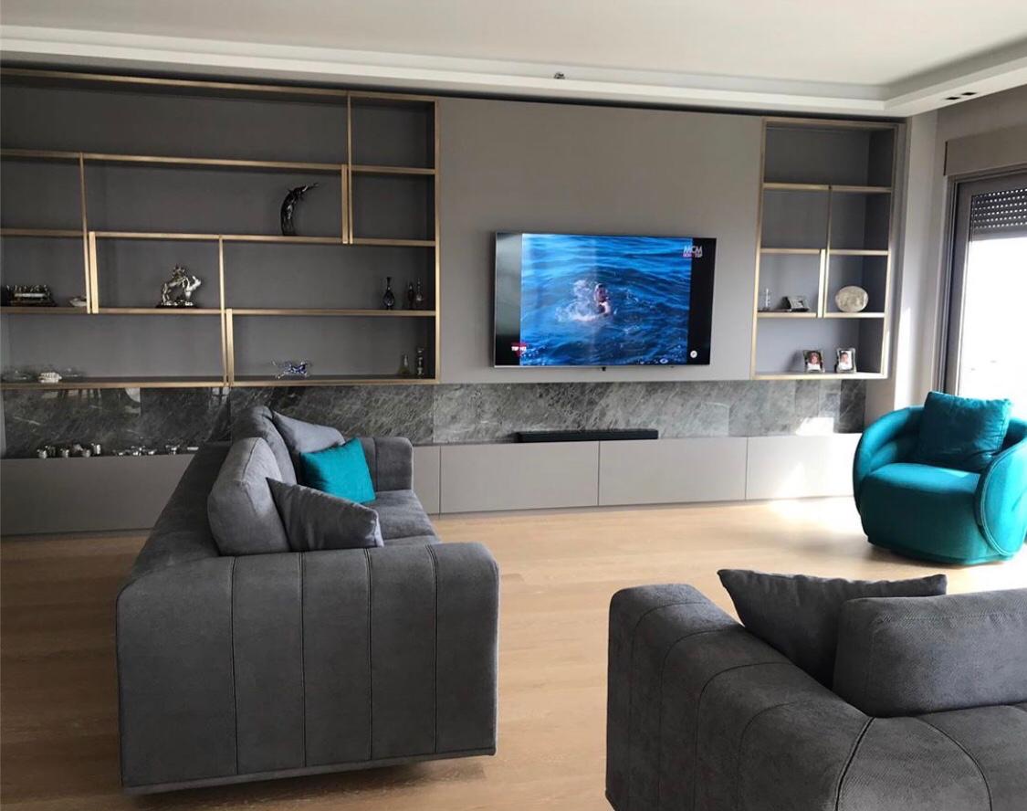 Modern living room featuring two gray sofas, a blue armchair, and a television mounted on a wall with built-in shelves. Decorative items are displayed on the shelves. The room has large windows and wooden flooring.