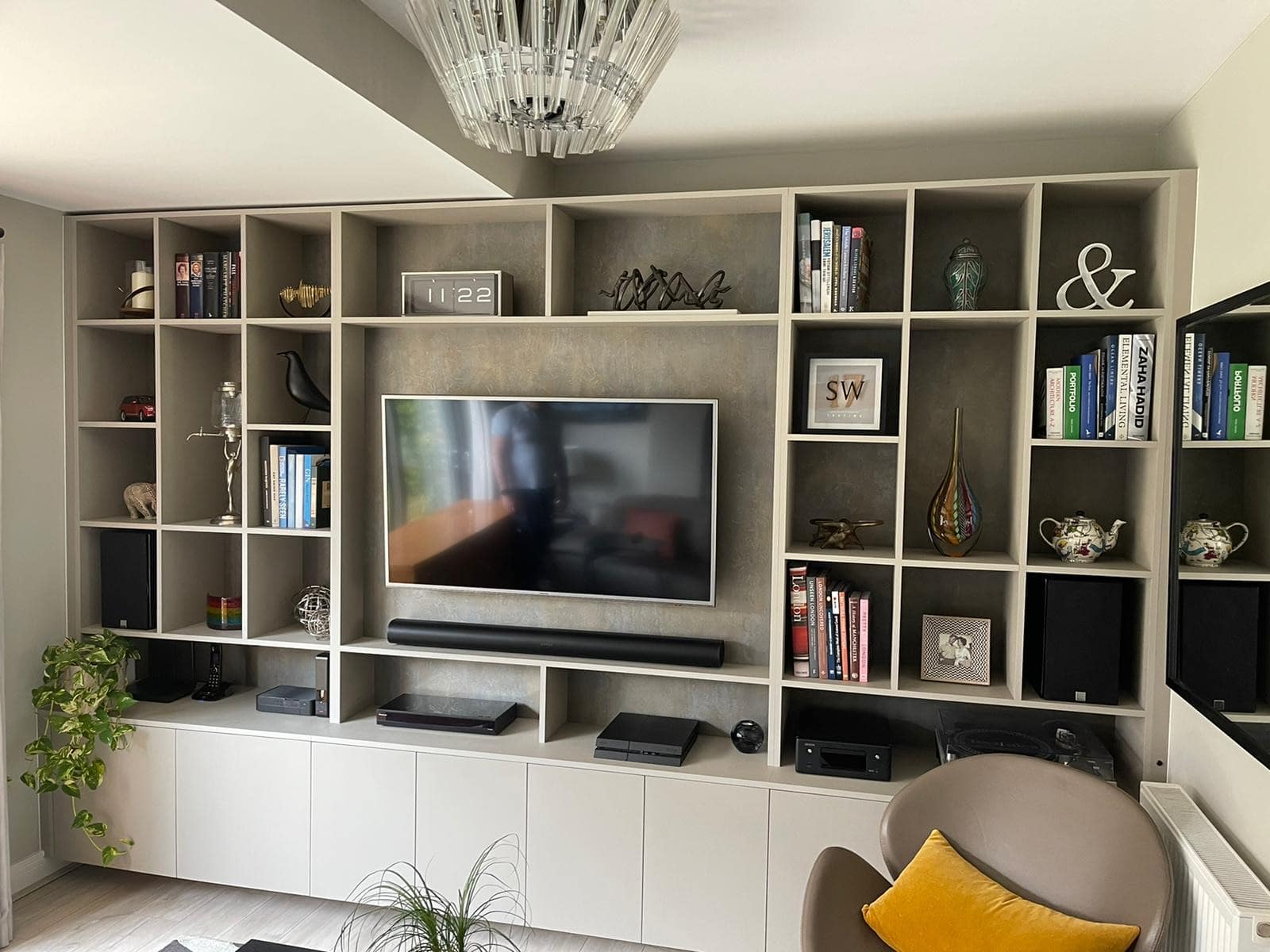 A modern living room features a wall-mounted TV surrounded by open shelving filled with books, decor items, plants, and electronics. The shelves are integrated into a sleek, minimalist entertainment unit with a mix of cubbies and closed storage.