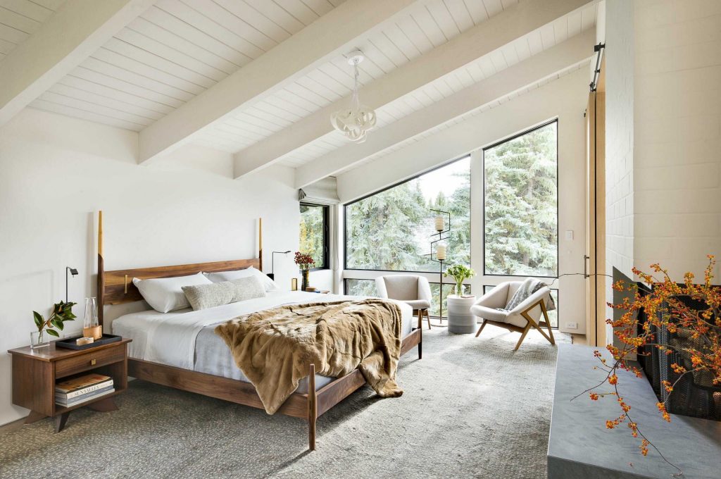 A bright, airy bedroom with a large bed, white linens, wooden accents, bespoke furniture including two chairs by the window, and a view of greenery outside. A chandelier hangs above, adding elegance to the room.
