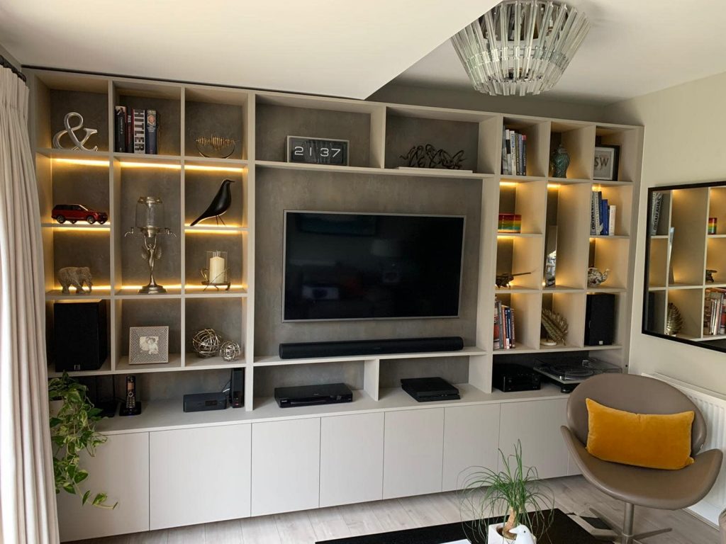 A modern living room featuring a large built-in wall unit with shelves containing books, decorative items, and a central flat-screen TV. There are spotlights in some compartments and a stylish chair in the corner. The room also includes fitted wardrobes that complement the overall décor.