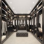 A luxurious and spacious fitted walk-in wardrobe boasts dark wood shelves and racks. Clothes, shoes, and accessories are neatly organized. An upholstered ottoman stands in the center of the room, which is brightly lit with recessed ceiling lights.