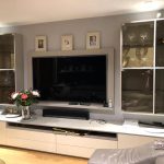 A modern living room featuring a large flat-screen TV mounted on a light grey wall. Flanking the TV are two glass-doored cabinets displaying various decorative items and trophies. A bouquet of flowers is placed on a light-colored cabinet below the TV.