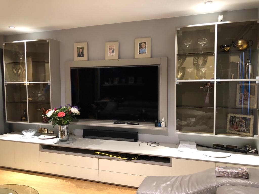 A modern living room featuring a large flat-screen TV mounted on a light grey wall. Flanking the TV are two glass-doored cabinets displaying various decorative items and trophies. A bouquet of flowers is placed on a light-colored cabinet below the TV.