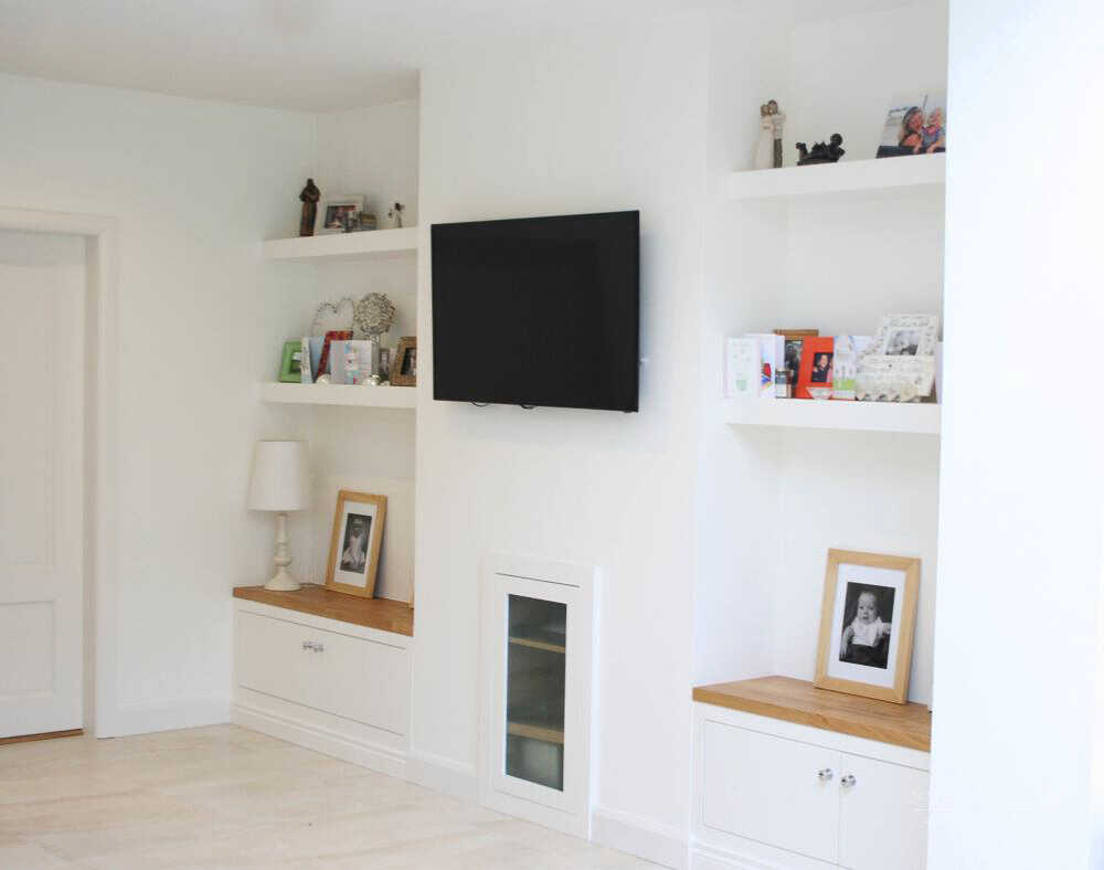 A minimalist living room features white walls, built-in shelves, and cabinets. A flat-screen TV is mounted above a small fireplace nestled in one of the alcoves. The shelves display various decorations, family photos, and a table lamp.