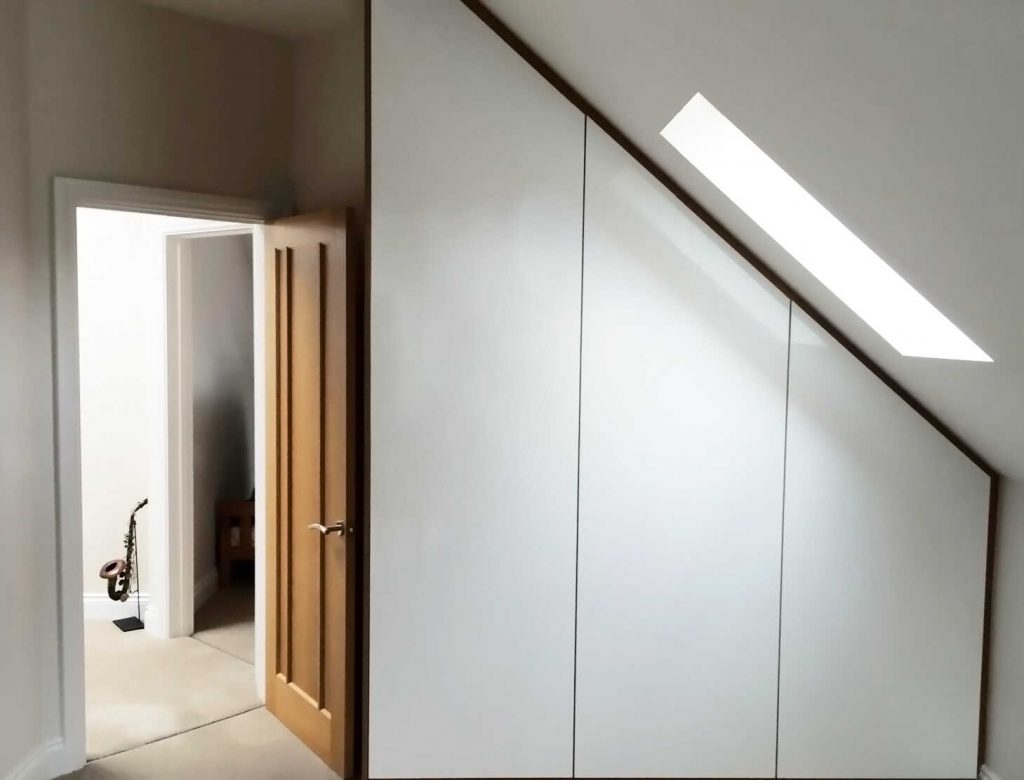 Modern, minimalist attic room featuring white fitted wardrobes under a sloped ceiling with a skylight. An open wooden door leads to another room.