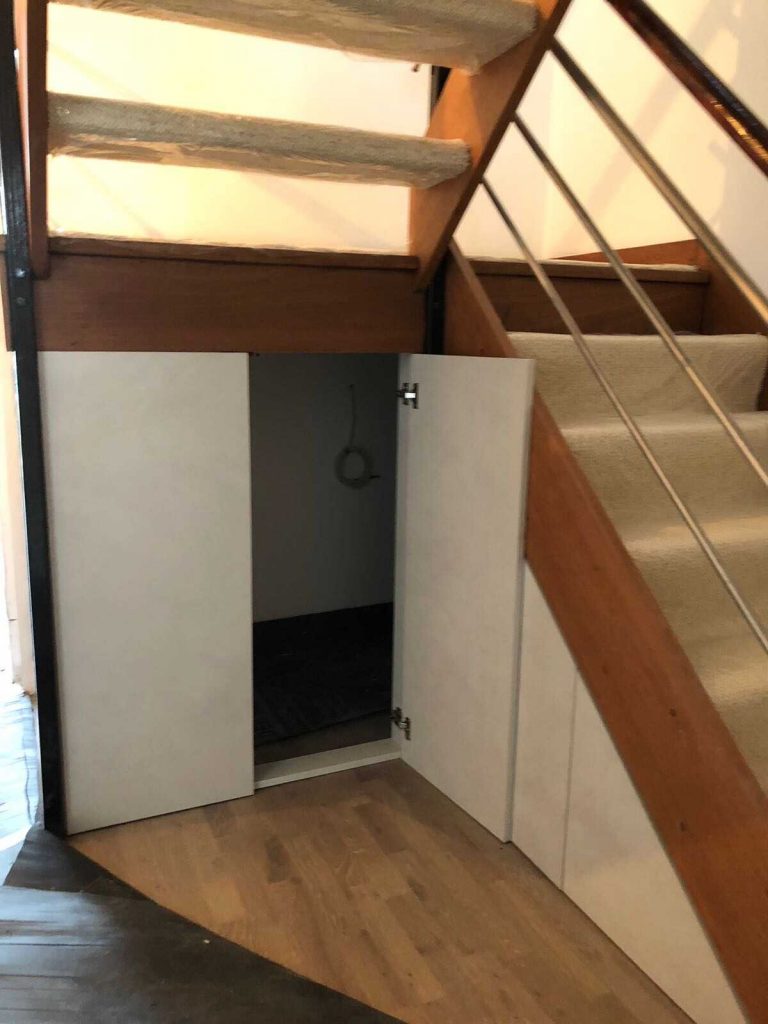 A small storage space under a staircase, accessible through a set of white double doors with bespoke furniture design, featuring a ring handle on one door. The staircase has wooden steps and a metal railing.