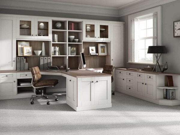 A spacious home office with a white L-shaped desk, brown leather office chair, and bespoke cabinet furniture filled with books and decor. A computer monitor sits on the desk. The room has large windows with shutters, gray carpet, and light gray walls.