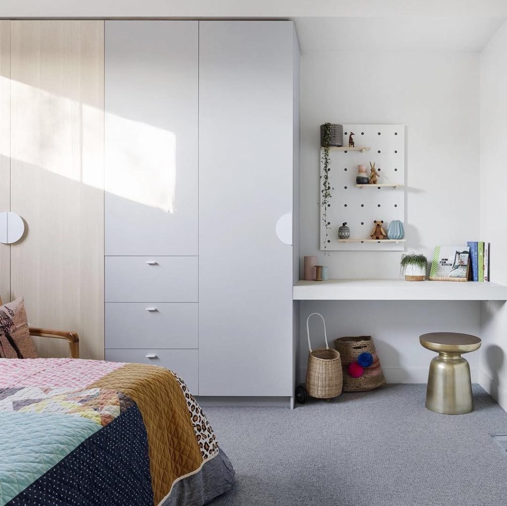 A modern, neat bedroom featuring a large bed with a colorful quilt, fitted wardrobes, and a small study area with a desk and shelving, bathed in natural sunlight.