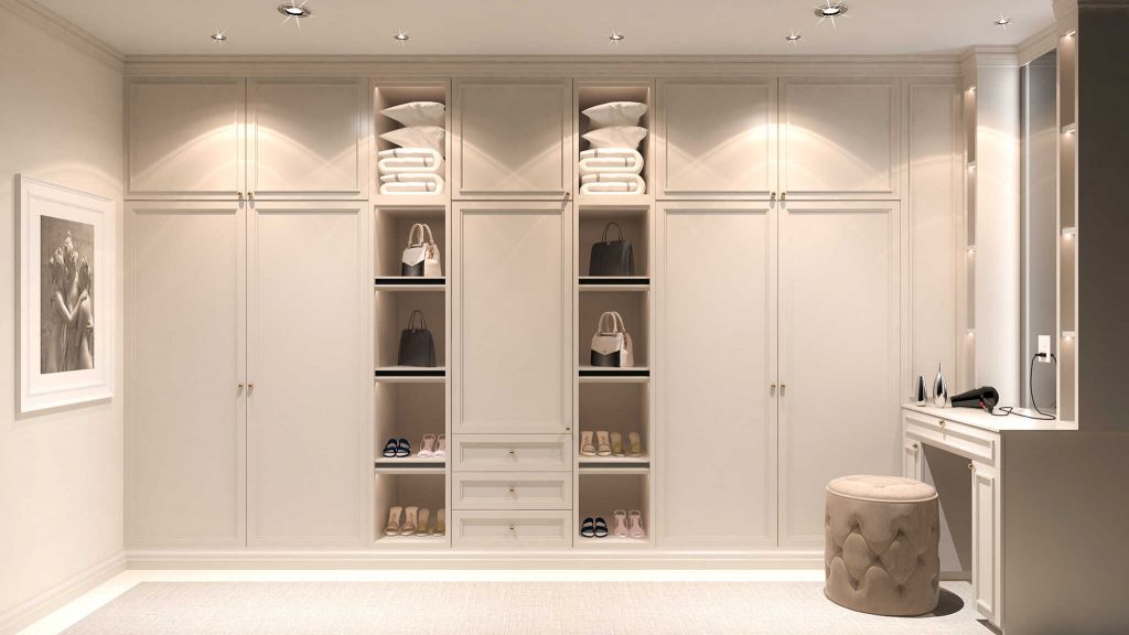 A modern walk-in wardrobe with white cabinetry, including shelves and closed compartments, neatly arranged with shoes and accessories, under soft lighting.
