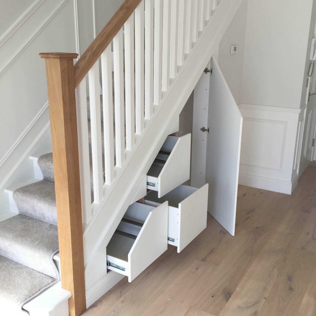 A staircase with built-in storage drawers underneath. The staircase is painted white with wooden railings and beige carpeting. Three pull-out drawers of different sizes are visible in the open storage area, revealing a smart use of space in the hallway.