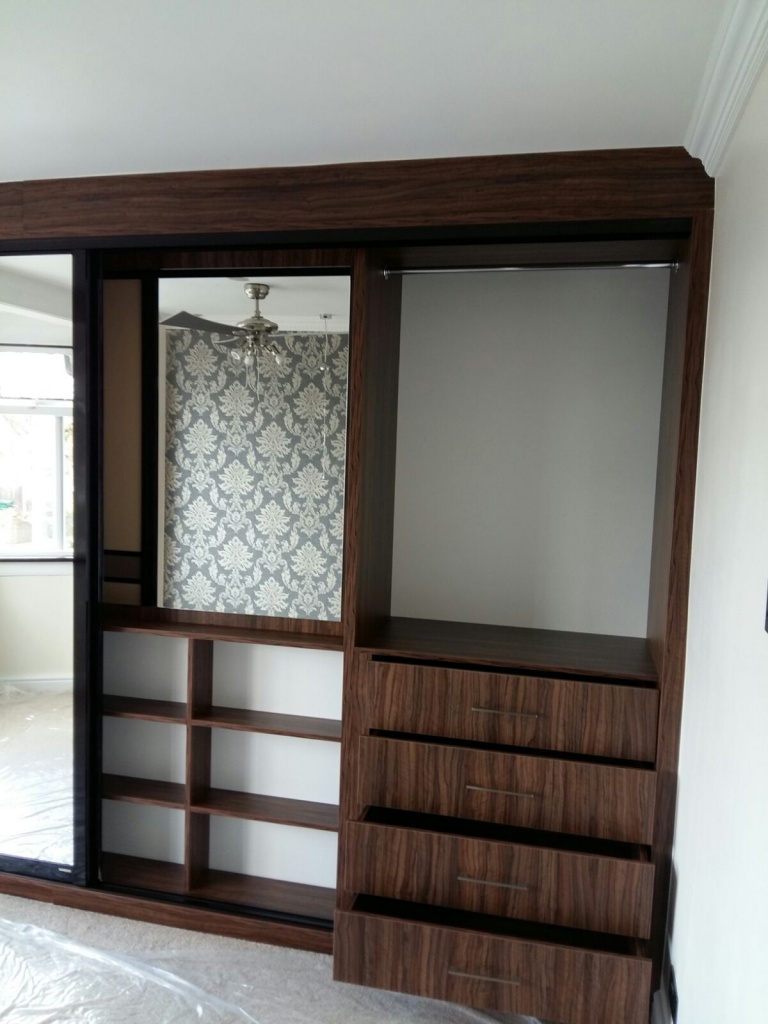A modern wooden fitted sliding door wardrobe with a built-in shelving unit and several drawers on the right. The wardrobe features sliding doors, one of which is a mirror. The background shows patterned wallpaper and a window, with a ceiling fan visible above.