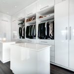 A modern, spacious walk-in closet featuring white fitted wardrobes and a central island, with neatly organized clothing hanging on both sides and dark wooden flooring.