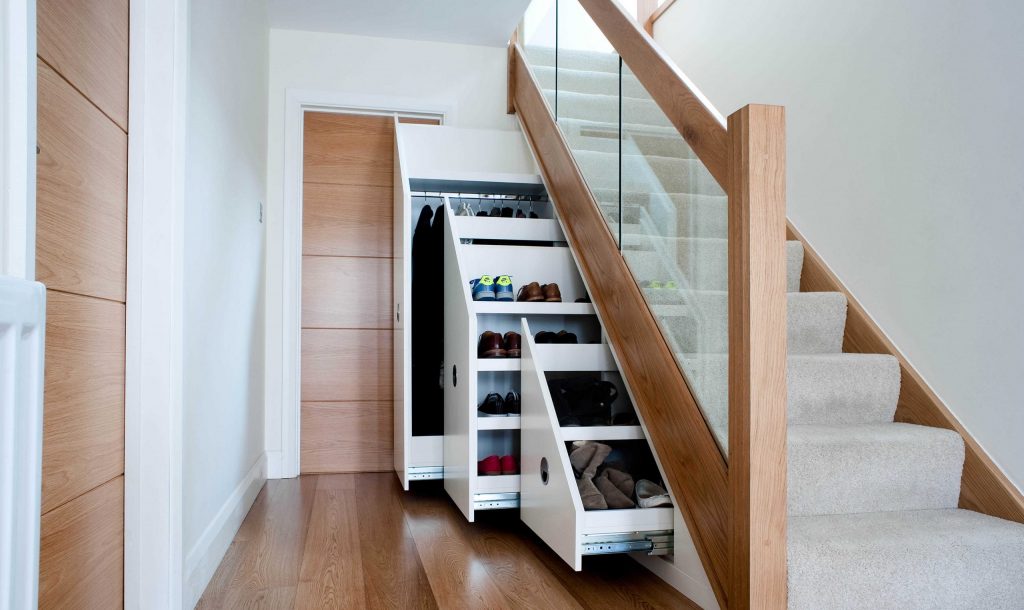 A modern staircase with built-in storage drawers underneath. The wooden and glass staircase has three pull-out drawers, each containing neatly organized shoes and other items. The space features a combination of light wood and neutral-colored walls.