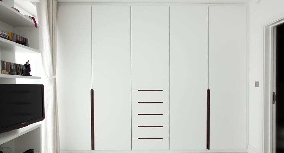 A sleek, modern wardrobe with white, flat-panel doors and a set of five central drawers in a minimalist room. To the left, bespoke furniture includes built-in white shelves with books and a television. Natural light streams in through a window curtain on the left side.