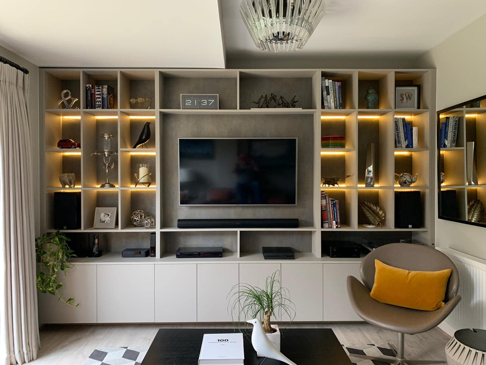 A modern living room features a large wall-mounted TV surrounded by backlit shelves displaying books, sculptures, and decor. Below the TV, there are closed cabinets. The room includes a cozy armchair with a yellow pillow, a ceiling light fixture, and a potted plant.