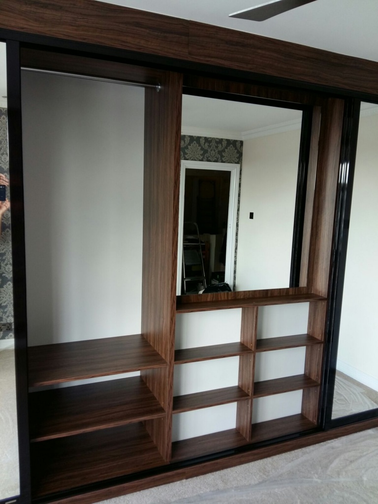 A modern walk-in wardrobe with sliding mirror doors, built-in dark wood shelving on one side, set in a room with patterned wallpaper visible through an open door.