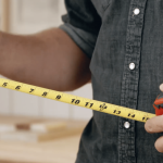 A person in a dark denim shirt uses a yellow measuring tape to measure a piece of wood for made-to-measure furniture. They hold a pencil in one hand, ready to mark measurements. The background is blurred, but it appears to be a workshop or woodworking space.