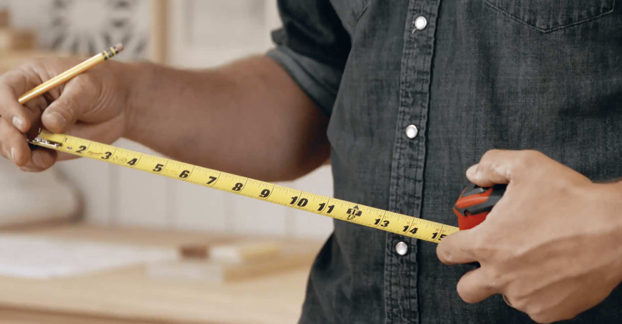 A person in a dark denim shirt uses a yellow measuring tape to measure a piece of wood for made-to-measure furniture. They hold a pencil in one hand, ready to mark measurements. The background is blurred, but it appears to be a workshop or woodworking space.